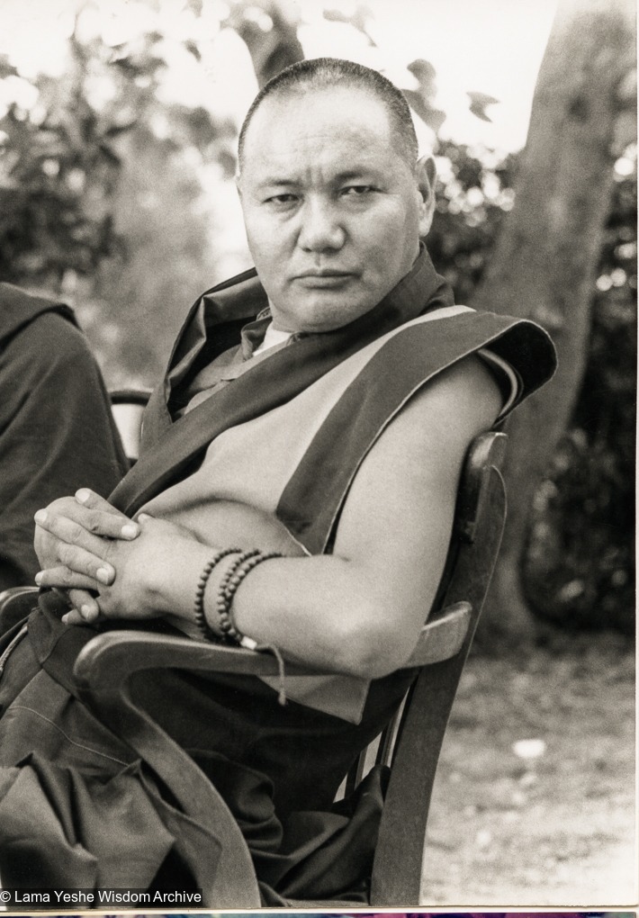 Lama Thubten Yeshe seated in a wooden chair outdoors, wearing traditional monastic robes.